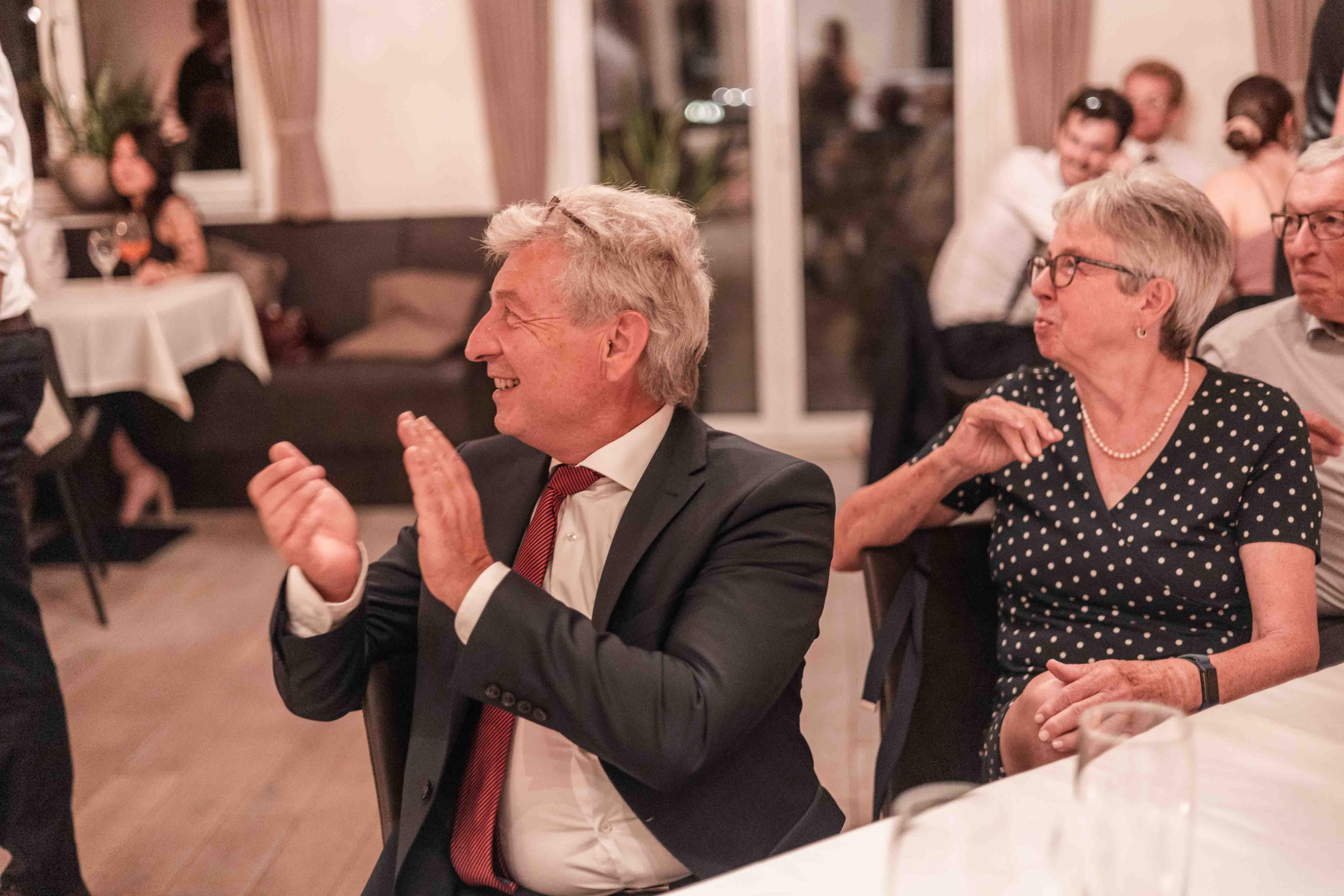 Hochzeitsfoto der Gäste in der Lokation an den Tischen sitzend - Das Abendprogramm ist gestartet und Gäste klatschen