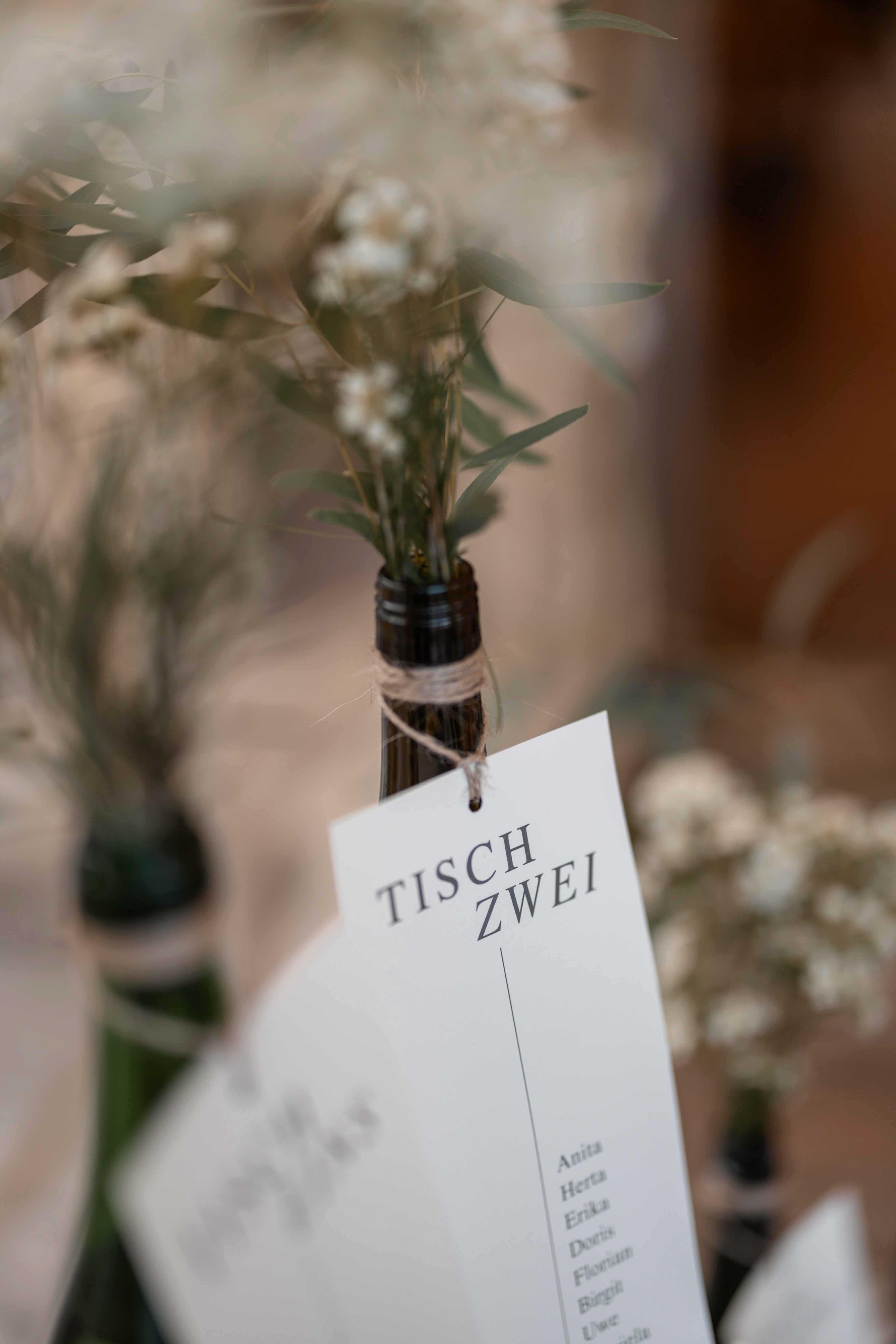 Fotografie der Dekoration - Eine Flasche mit Blumen dekoriert und einem Zettel auf dem Steht 'Tisch Zwei'