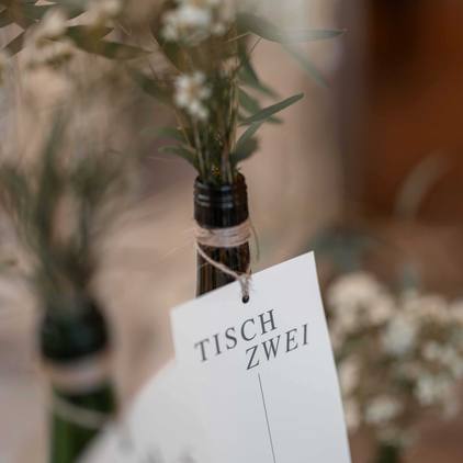 Fotografie der Dekoration - Eine Flasche mit Blumen dekoriert und einem Zettel auf dem Steht 'Tisch Zwei'